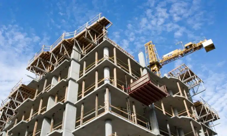Low-angle view of a multi-story concrete building under construction with a yellow tower crane and wooden scaffolding against a bright blue sky.