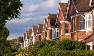 A long perspective view of red brick semi-detached houses with gabled roofs and bay windows under a blue sky with soft clouds.