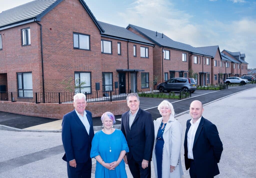 Five officials stand before newly completed brick terraced homes in Liverpool, part of the city&rsquo;s &pound;700 million social housing investment. The modern design features dark grey roofs, black-framed windows, and accessible frontage  symbolizing the delivery phase of the 63,000-home regional housing pipeline.