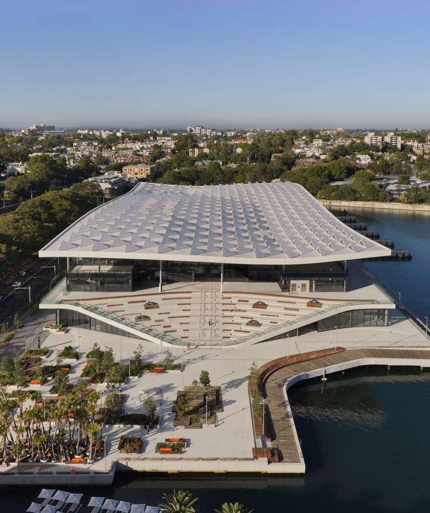 Aerial view of the Sydney Fish Market at Blackwattle Bay, showcasing its undulating roof canopy, terraced public plazas, and waterfront integration with urban greenery.