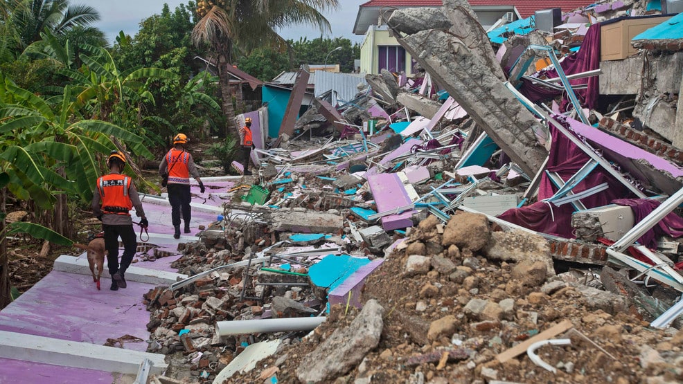 Rescue workers and a sniffer dog navigate debris after the Talud Islands earthquake 2026, with collapsed structures and exposed building materials visible amid tropical vegetation.
