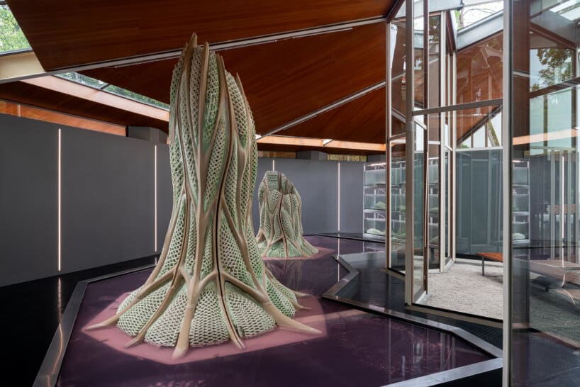 Living Building prototype with bio-integrated 3D-printed structure, showcasing carbon-absorbing microorganism-infused facade in exhibition setting under timber canopy.