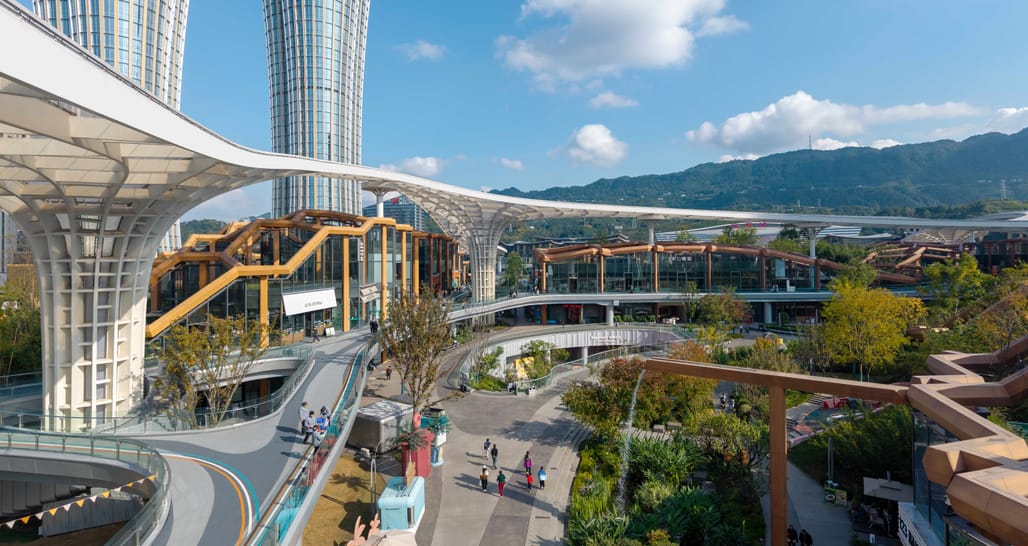 Pedestrian walkways weave through the central park of the Pelli Clarke Partners bamboo inspired Yibin transit district, with modern, nature integrated buildings under a sweeping white canopy.