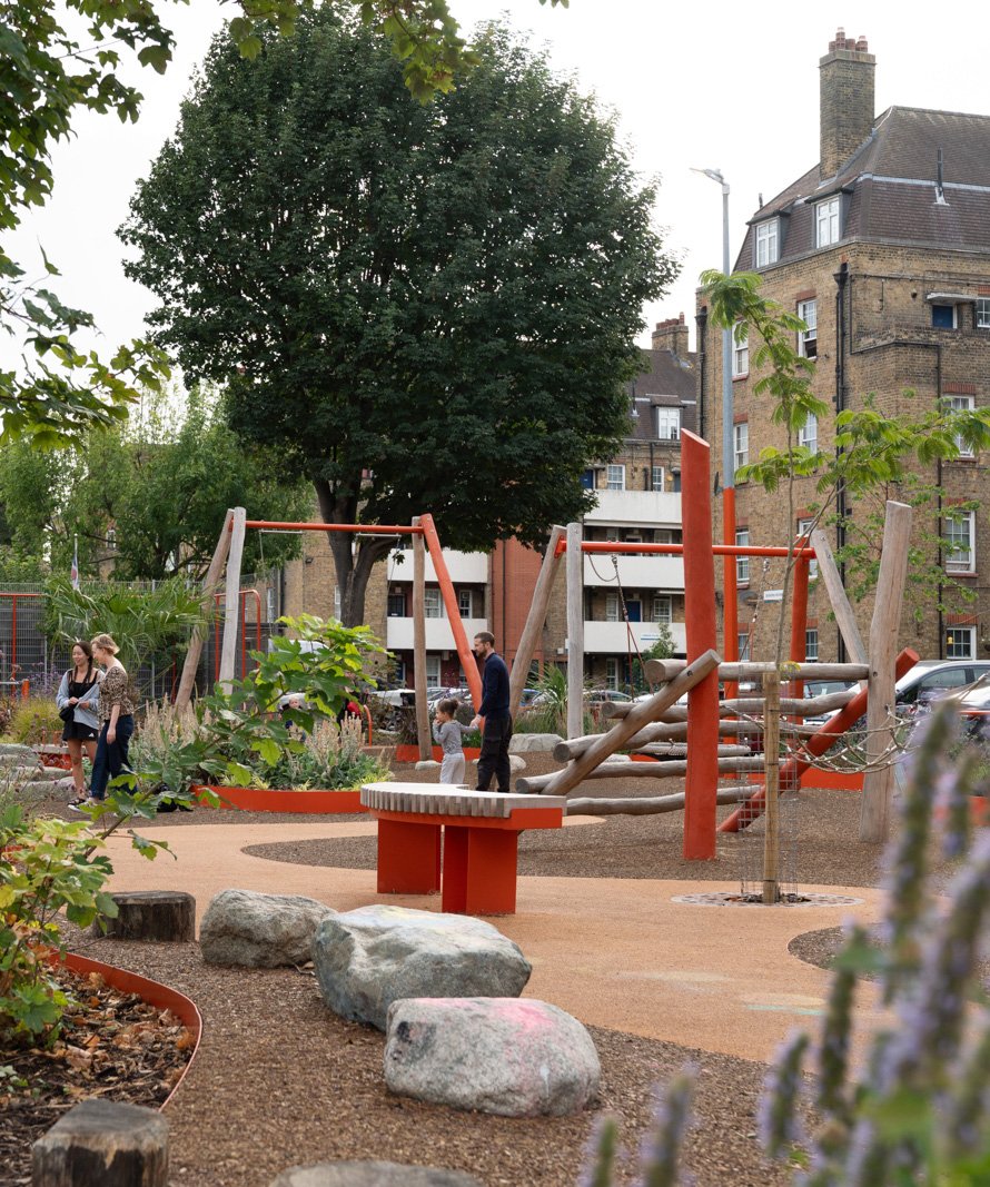 A father and child interacting in the redesigned Frederick Adventure Playground, with its open design featuring naturalistic landscaping and orange-accented play equipment.