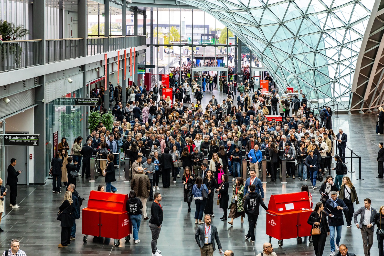 A sleek, modern design kiosk with bold red lettering at the Salone del Mobile 2026, illustrating the event's updated branding and visual identity.