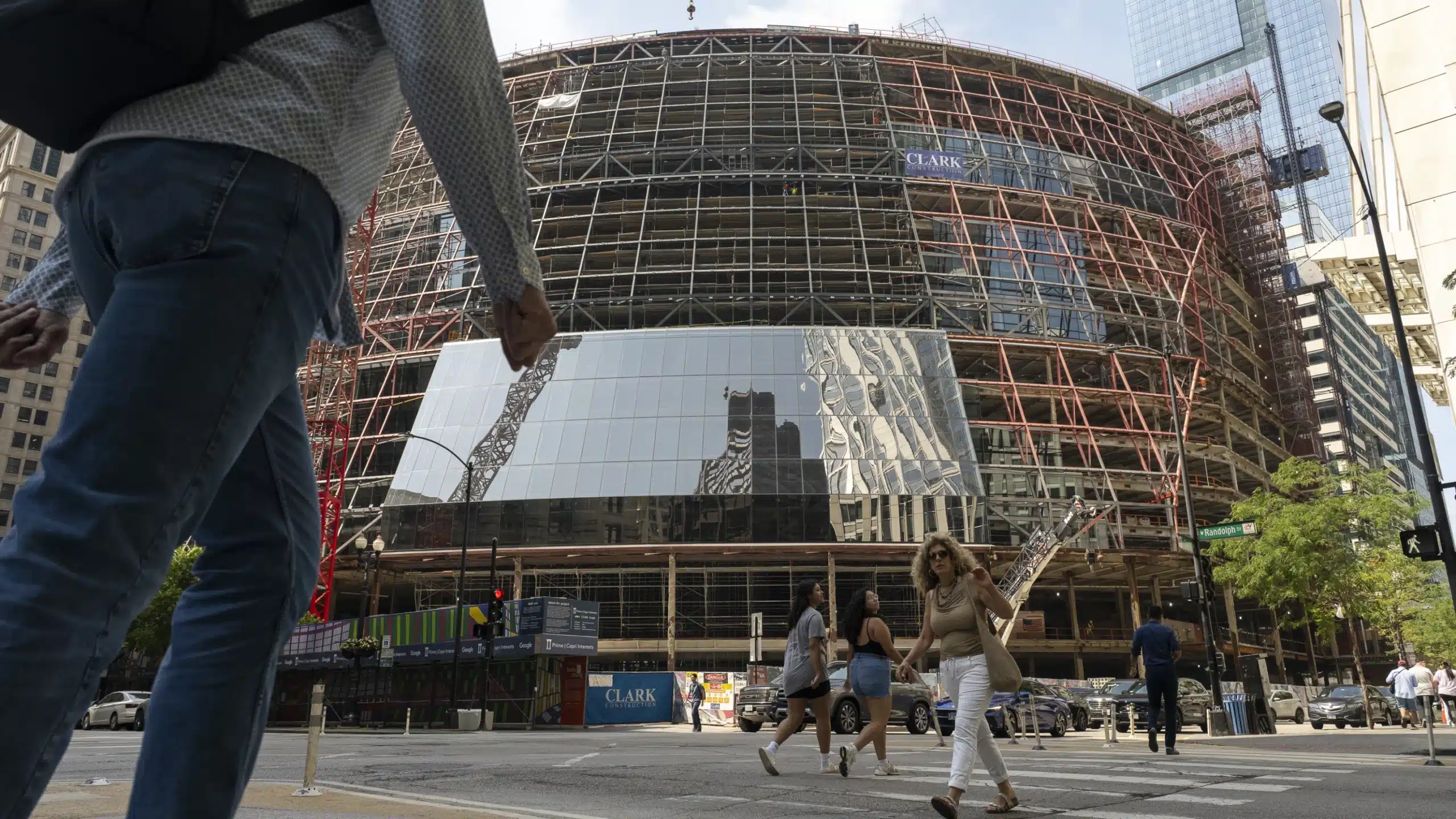 Pedestrians walk past the partially clad Google Chicago HQ under construction, with scaffolding and steel framework visible against the urban skyline.