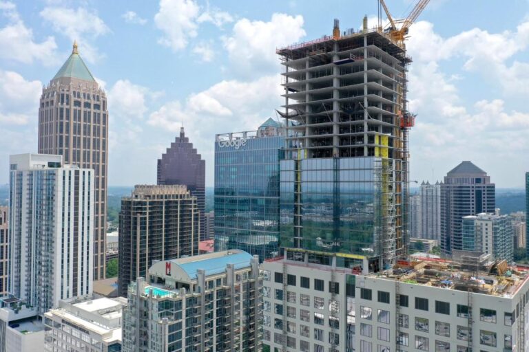 Atlanta mixed-use tower under construction, rising above Midtown skyline with exposed concrete frame and crane against blue sky.