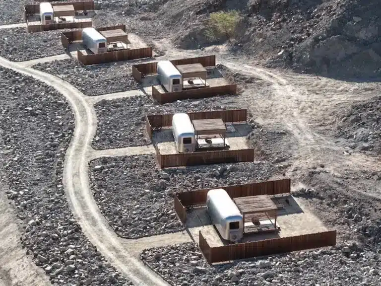 Aerial view of Nomad Kalba’s off-grid accommodation trailers nestled in rocky mountain terrain, each enclosed by low wooden fences and connected by winding dirt paths — environmental reuse applied to hospitality infrastructure.
