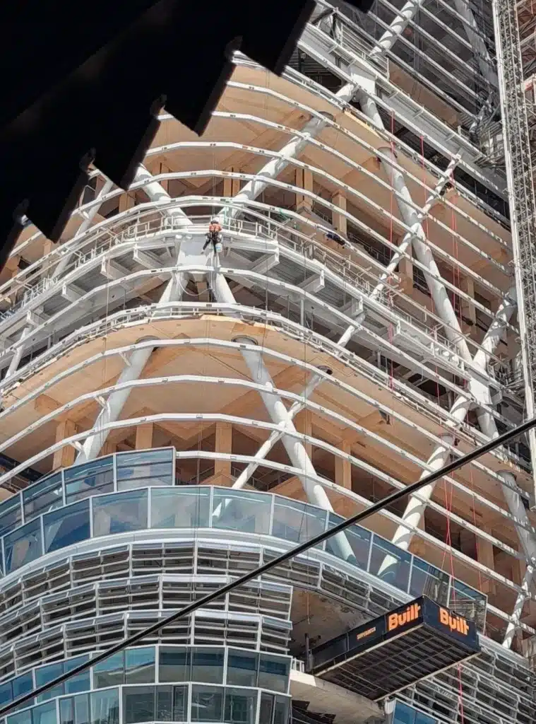 A construction worker suspended on the curved facade of a hybrid timber tower under development, showcasing exposed mass timber floors and white steel exoskeleton.