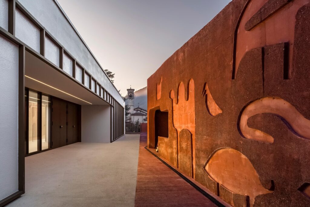 Modern architectural design courtyard with textured orange wall and linear building facade, church tower visible in distance.
