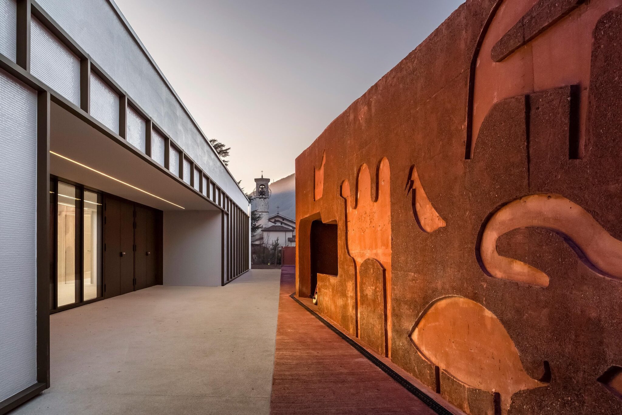 Modern architectural design courtyard with textured orange wall and linear building facade, church tower visible in distance.