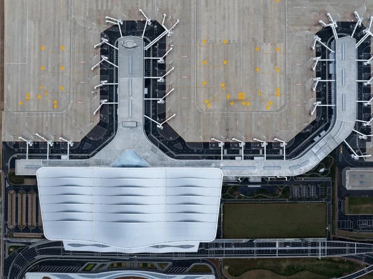 Aerial view of Yantai Terminal 2 showing its E-shaped layout, undulating roof, and jetways aligned with the runway. Terrain-responsive design minimizes excavation.
