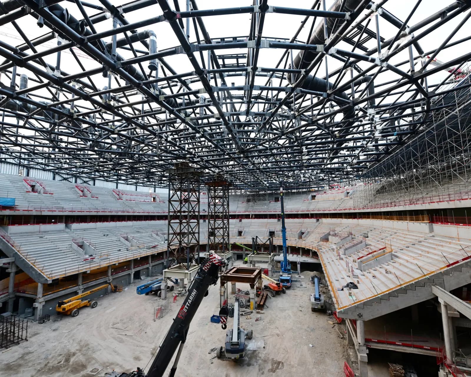 Interior view of a large circular arena under construction, showing exposed steel trusses and incomplete concrete seating tiers. Arena del ghiaccio di Milano