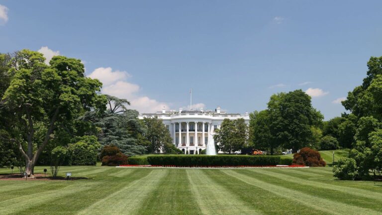 Frontal view of the under clear sky, framed by manicured lawns and mature trees, with fountain visible at center.
