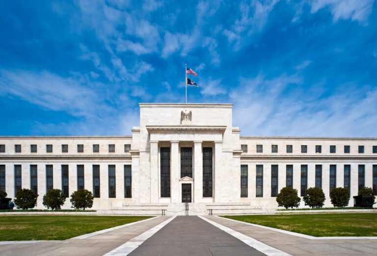 Frontal view of the Eccles Building, headquarters of the Federal Reserve in Washington, D.C., showcasing its neoclassical façade, central portico with fluted columns, carved eagle emblem, and American flag against a blue sky symbolizing institutional authority and architectural preservation.