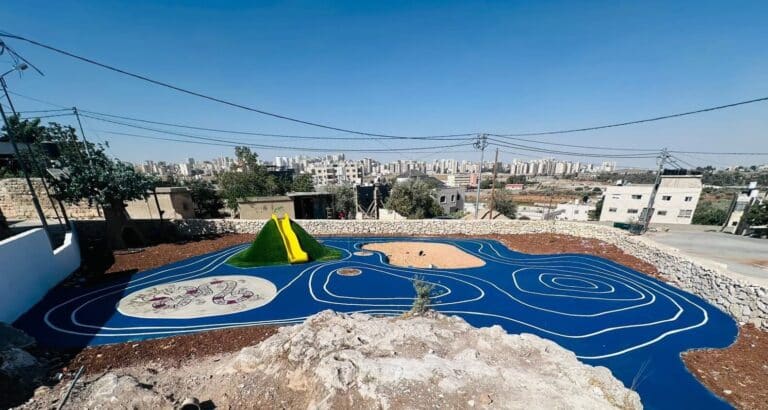 Blue topographic playground with white contour lines and artificial green mound, set against urban skyline in Qalandiya, Palestine.