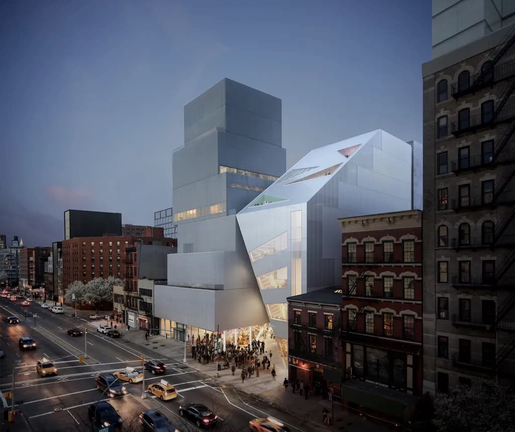Aerial evening view of the Bowery Contemporary Art Museum expansion in New York, showcasing its geometric glass and metal façade integrated with historic brick buildings on a bustling urban street.