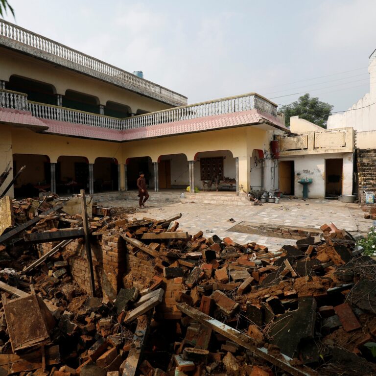 A man walks through the courtyard of a building in Kashmir damaged by the earthquake, with a large pile of brick rubble in the foreground.
