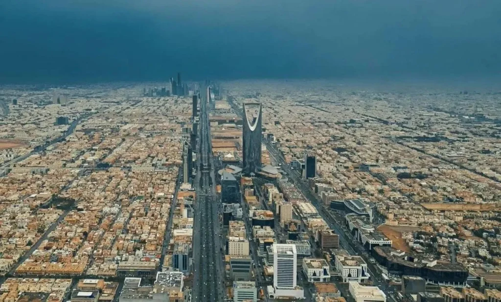 An expansive aerial view of Riyadh, Saudi Arabia, featuring the iconic Kingdom Centre skyscraper and a long highway stretching toward a distant city horizon under a dramatic, cloudy sky.