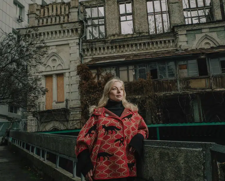A blonde woman wearing a red and black patterned coat with fox motifs standing in front of a crumbling, abandoned neo-Gothic stone building under an overcast sky.