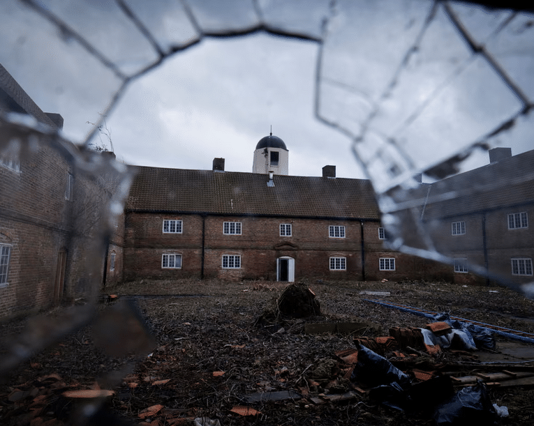 A view through a large jagged hole in a broken glass window looking onto a derelict two-story brick building and a cluttered, overgrown courtyard under a gloomy sky.