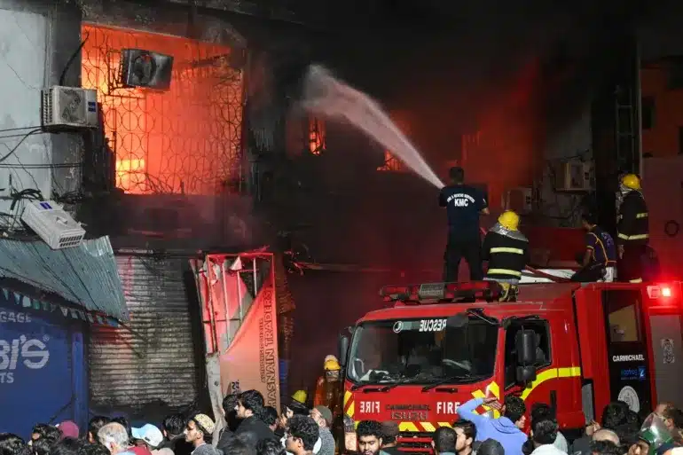 Firefighters battle intense flames engulfing a multi-storey commercial building in downtown Karachi during the January 18, 2026 urban shopping center fire, as crowds observe from street level.