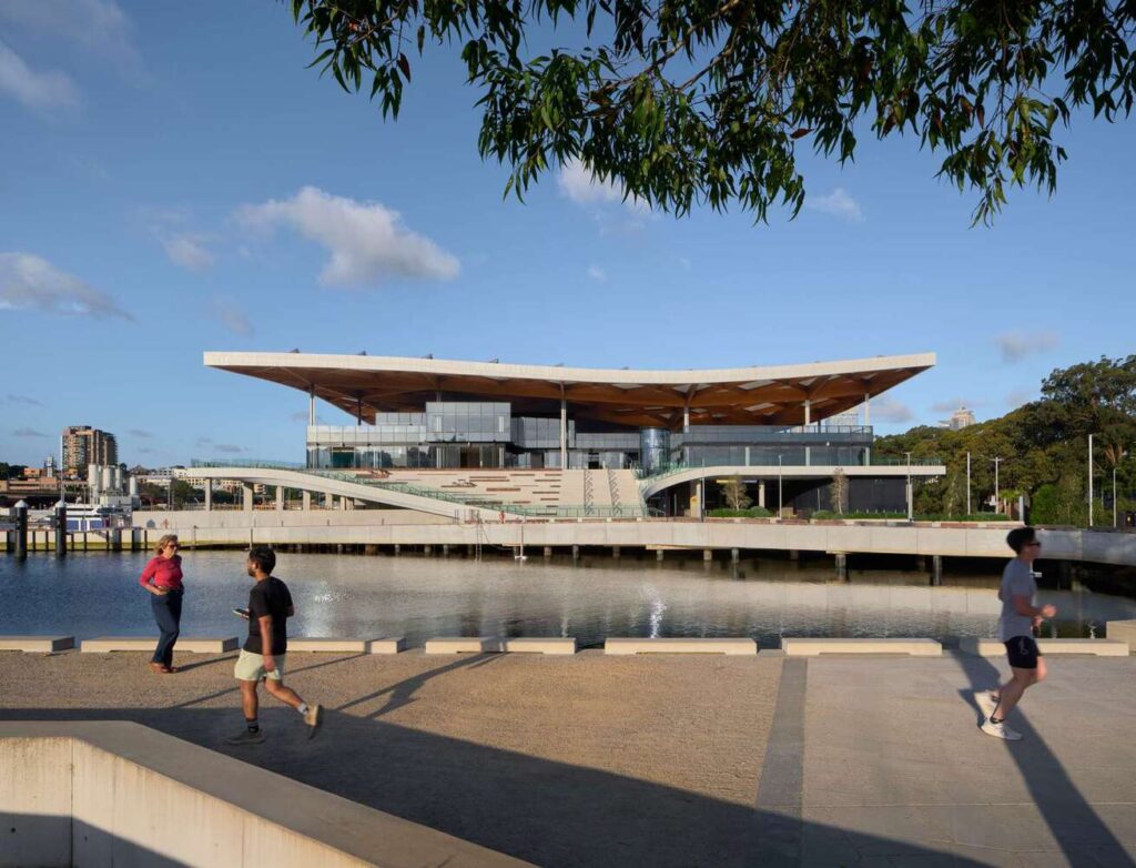 Sydney Fish Market at Blackwattle Bay, viewed from the waterfront promenade, with pedestrians walking along the edge and its cantilevered roof extending over glass facades.