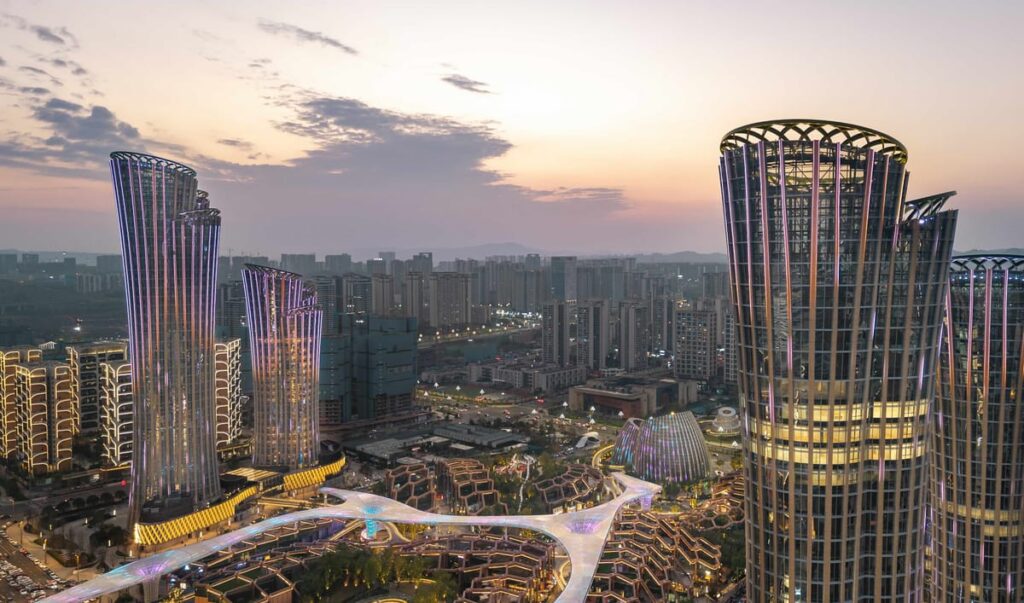 The illuminated bamboo stalk inspired towers of the Yibin High Speed Railway Station development dominate the city skyline at dusk, showcasing their curved silhouettes and vertically articulated facades