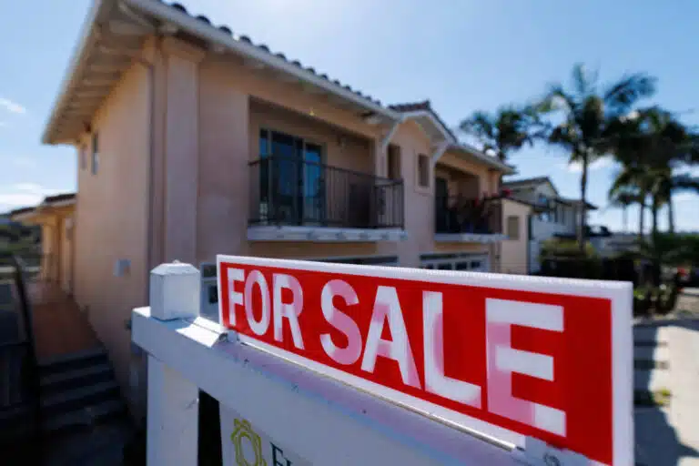 Close-up of a red and white "For Sale" sign in front of a blurred two-story residential house under a clear blue sky.