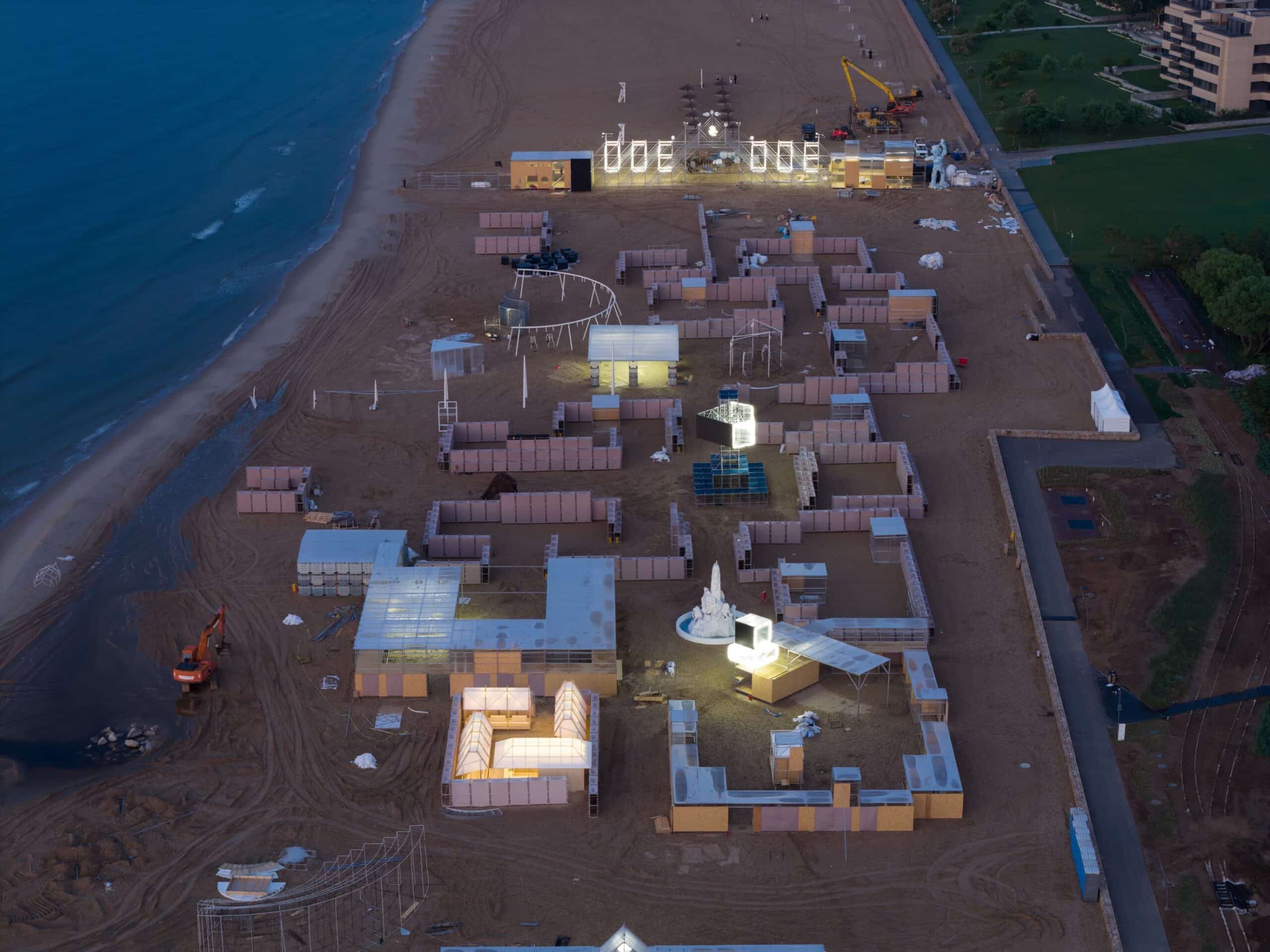 Aerial view of a modular temporary pavilion with white fabric roofs and wooden frames, arranged around a central courtyard on sandy terrain exemplifying ephemeral architecture.