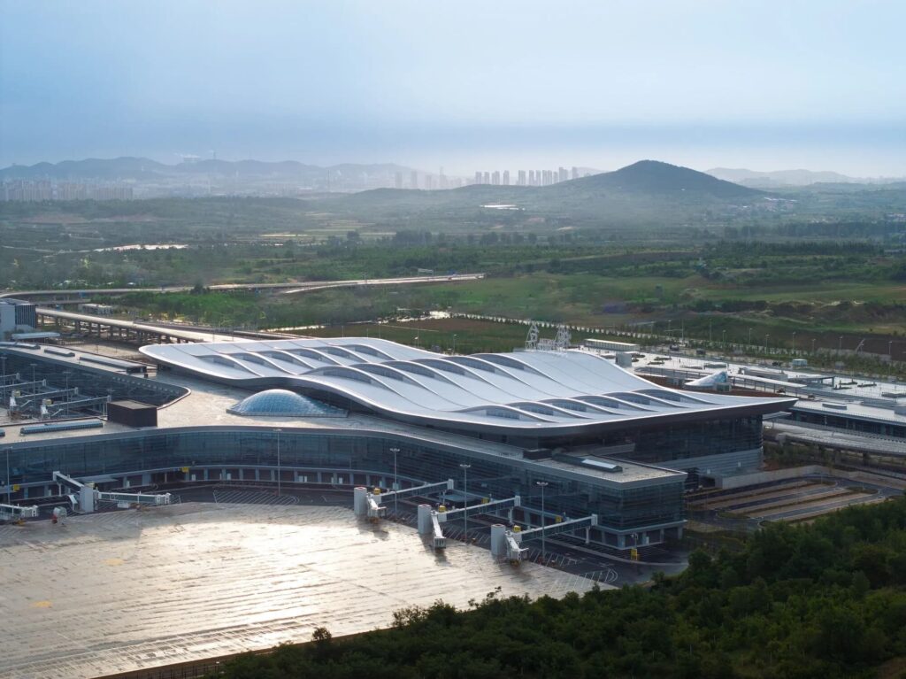Yantai Terminal 2 aerial view showing its wavy roof structure, glass facade, and integration with surrounding hills and urban skyline. Terrain-responsive airport infrastructure design.