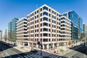 A mid-century modern office building in downtown Washington, featuring a grid-like façade of concrete and glass, situated at a street corner with pedestrian crossings and adjacent contemporary glass structures under a clear blue sky.