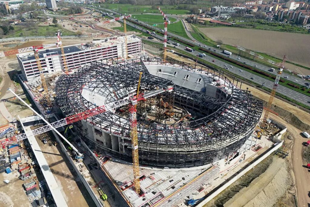 منظر جوي لصالة هوكي الجليد الأولمبية في ميلانو 2026 قيد الإنشاء في سانتا جوليـا، يُظهر هيكلها الدائري الفولاذي، والرافعات المحيطة، والبنية التحتية الحضرية المجاورة قبل انطلاق الألعاب الشتوية.