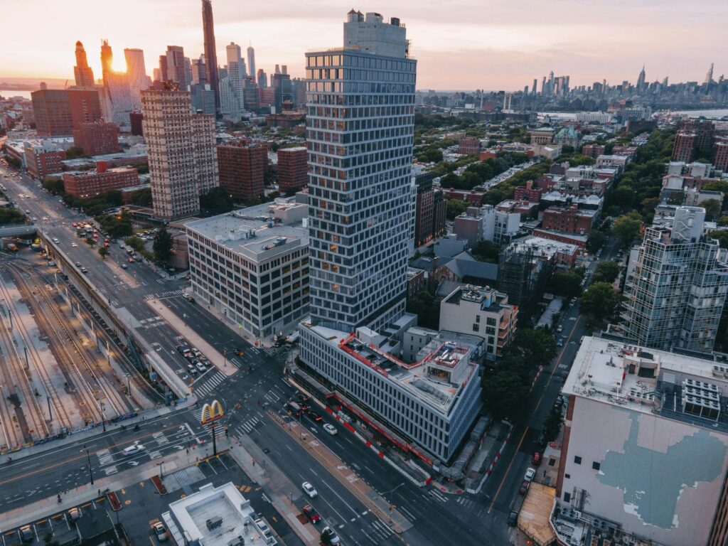 Aerial view of a modern mixed-use tower in Brooklyn at sunset, surrounded by urban fabric, elevated train tracks, and the Manhattan skyline in the background.