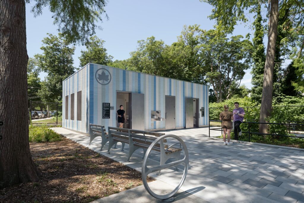 Exterior view of the AIANY Award-winning Staten Island public restroom at Lopez Playground, featuring blue-and-white striped tile cladding, accessible entrances, and integrated benches under mature trees.