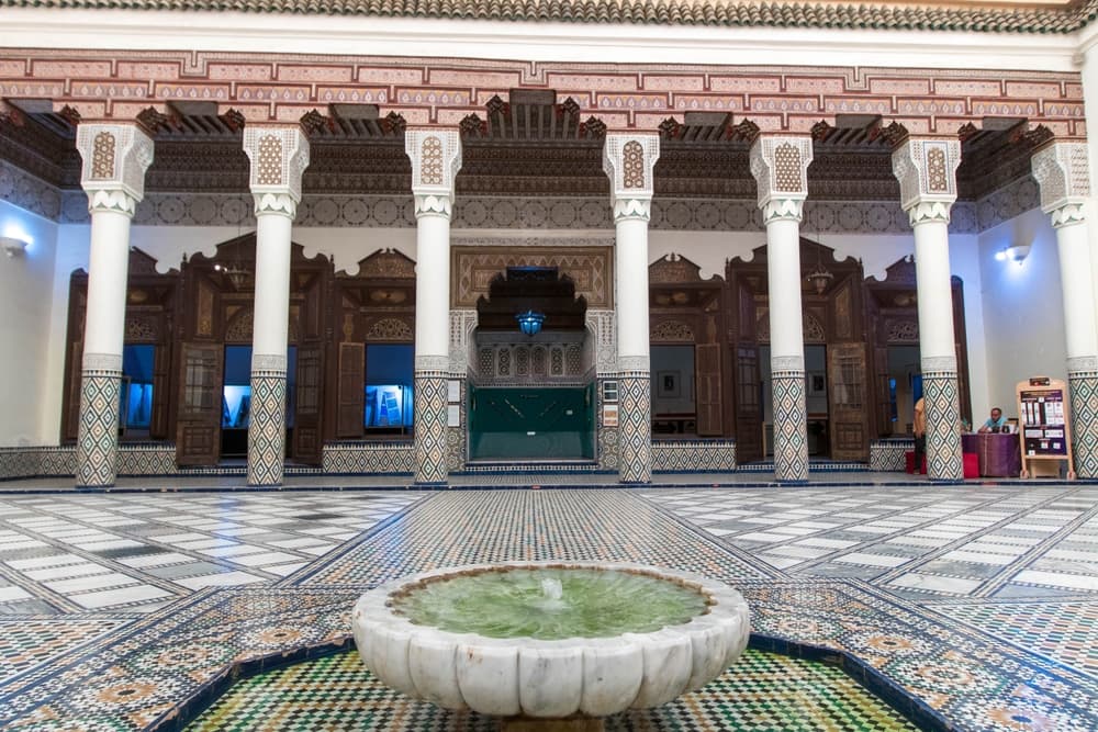 Grand courtyard of a historic Moroccan palace or public bathhouse in Fez or Marrakech, featuring ornate zellige tilework, white columns with carved capitals, and a central marble fountain  exemplifying Islamic architectural principles of symmetry, water as social anchor, and passive climate control.
