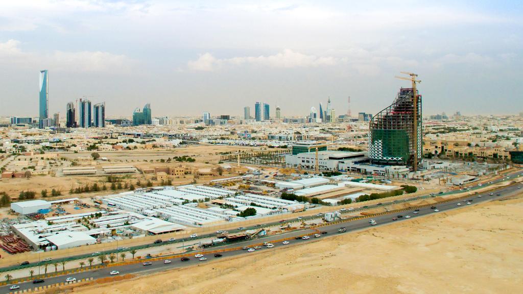 An aerial panoramic view of the Riyadh skyline featuring the Kingdom Centre and Al Faisaliyah Center, with a large modern building under construction in the foreground and a desert landscape under a cloudy sky.
