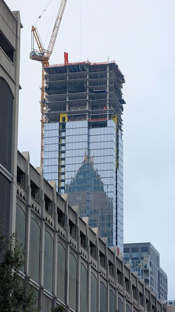 Construction crane beside partially clad , reflecting historic building in its glass facade under overcast sky.