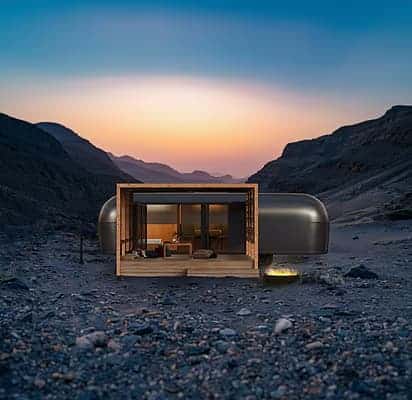 وحدة إقامة نوماد كلباء عند الغروب، تتميز بهيكل معدني مع إطار خشبي مفتوح وإضاءة داخلية دافئة، وسط تضاريس جبلية  إعادة الاستخدام البيئي في تصميم الضيافة خارج الشبكة.