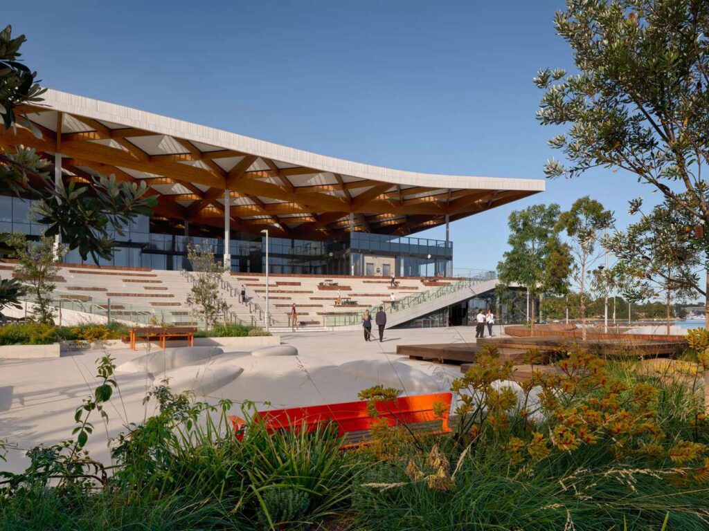 Sydney Fish Market at Blackwattle Bay, featuring its curved timber roof structure, tiered public seating, and landscaped foreground with native plantings under clear daylight.