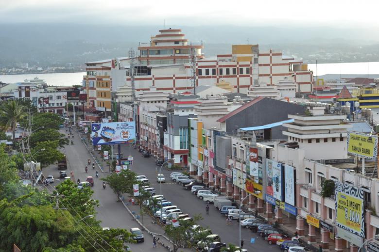 Aerial view of Manado city center showing commercial streets lined with multi-story buildings, parked vehicles, and signage, with coastal hills visible in the background under overcast skies.