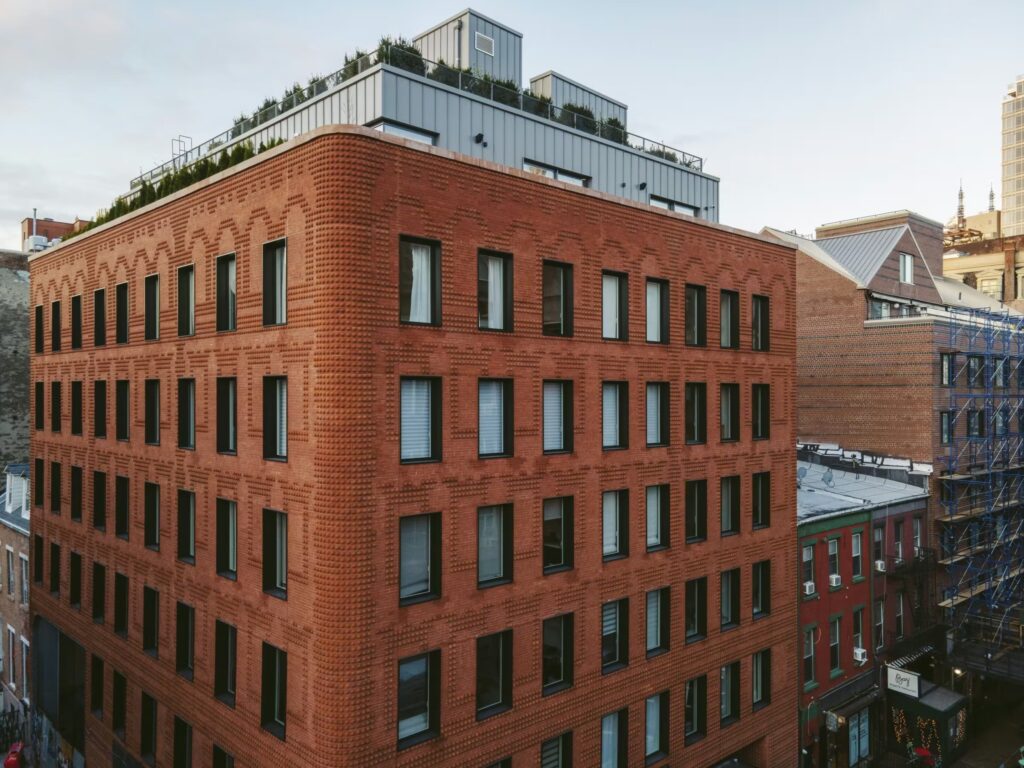 Modern brick residential building in New York with sculpted fa&ccedil;ade pattern, black-framed windows, and green rooftop terrace under overcast sky.
Architectural Innovation