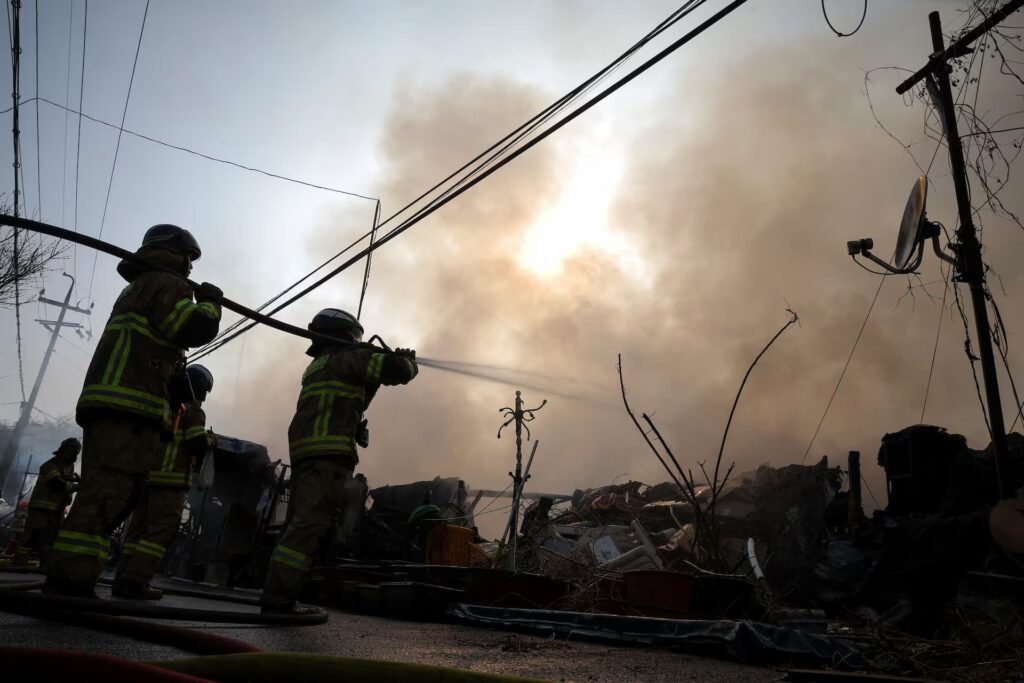 Firefighters battle the January 2026 in Guryong Village, Seoul, silhouetted against thick smoke and tangled utility lines &mdash; a scene reflecting the challenges of emergency response in densely packed informal settlements.