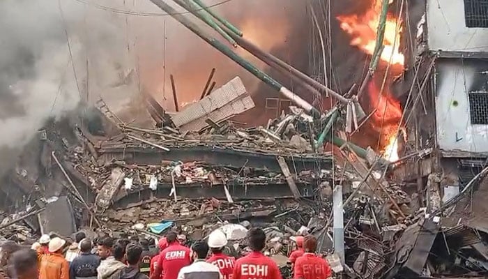 Collapsed debris and active flames at the site of the January 18, 2026 urban shopping center fire in downtown Karachi, with rescue workers from EDHI observing structural damage amid smoke and rubble.