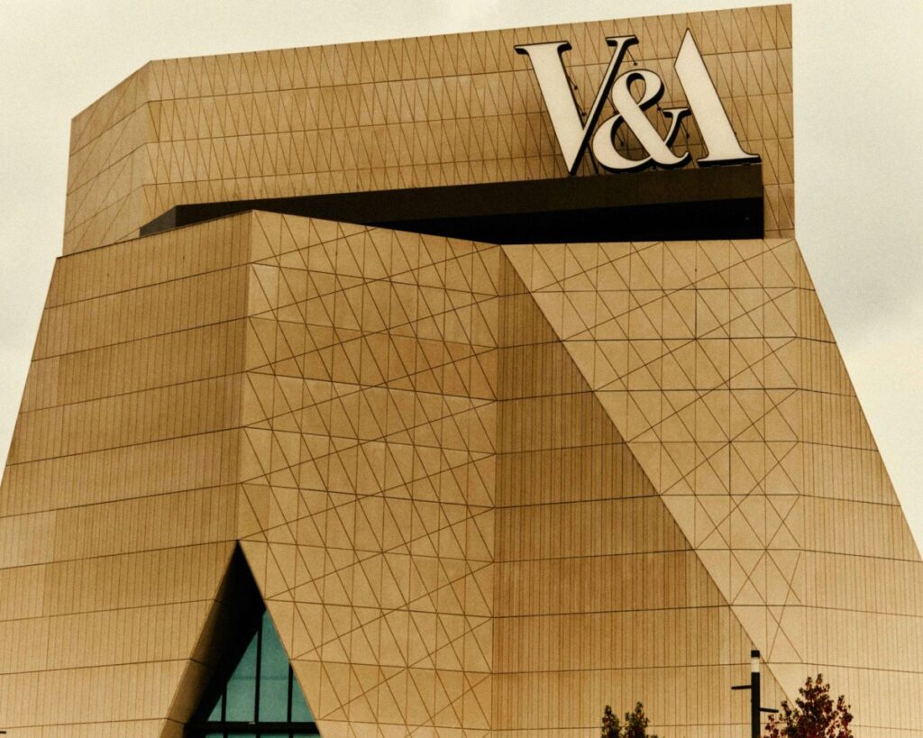 A close-up photograph of the V&A East Museum's geometric facade pattern and the prominent V&A logo at the top of the building