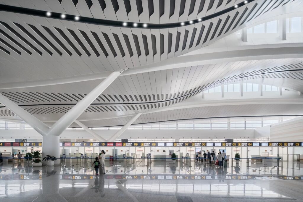 Yantai Terminal 2 interior showing sculptural ceiling with linear voids, structural columns, and security checkpoints. Natural light floods the space through high windows.