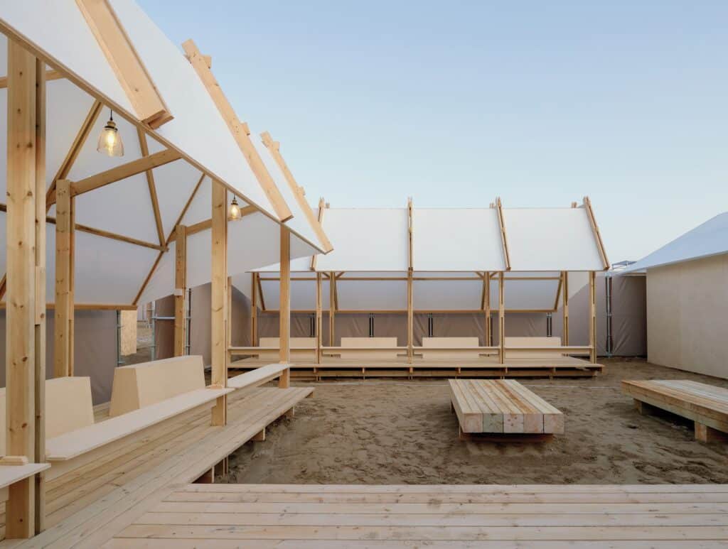 منظر داخلي لهيكل مؤقت يعرض عوارض خشبية مكشوفة وأسقف نسيجية بيضاء، مع مقاعد خشبية مدمجة وإضاءة معلقة &mdash; يجسد مفهوم العمارة الزائلة.