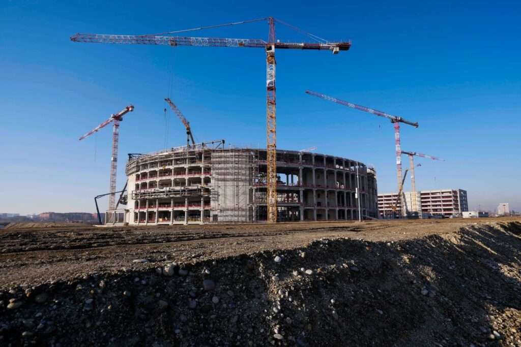 Ground-level view of the Milan 2026 Olympic Ice Hockey Arena under construction, featuring its multi-tiered concrete frame, scaffolding, and towering cranes against a clear blue sky.