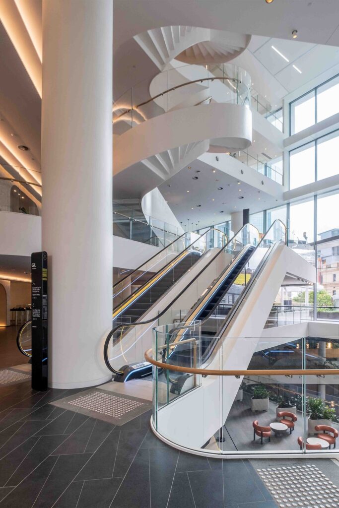 A multi-level interior atrium in a Sydney office building featuring a sculptural white staircase and glass-enclosed escalators, designed to guide movement through spatial layering.