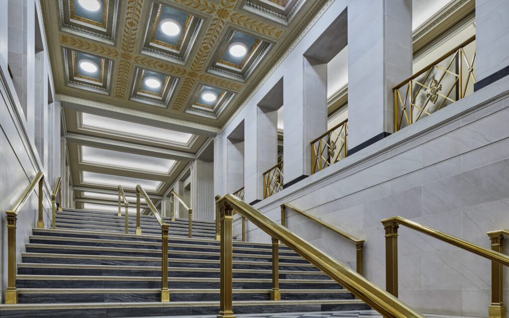 Grand interior staircase of the Federal Reserve&rsquo;s Eccles Building in Washington, D.C., featuring polished dark marble steps, gilded bronze railings, and a coffered ceiling with ornamental gold leaf detailing &mdash; exemplifying neoclassical grandeur preserved through 2026 renovation.